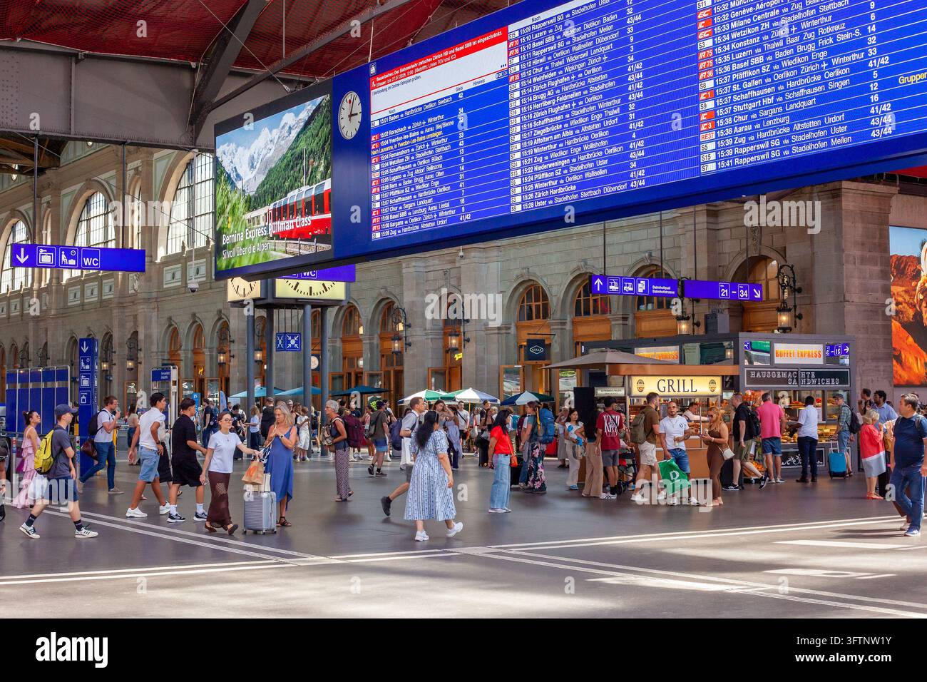 ZURICH, SUISSE - 5 JUILLET 2026 : Passagers marchant sous le panneau d'horaires d'arrivée et de départ bleu à la gare principale de Zurich Banque D'Images