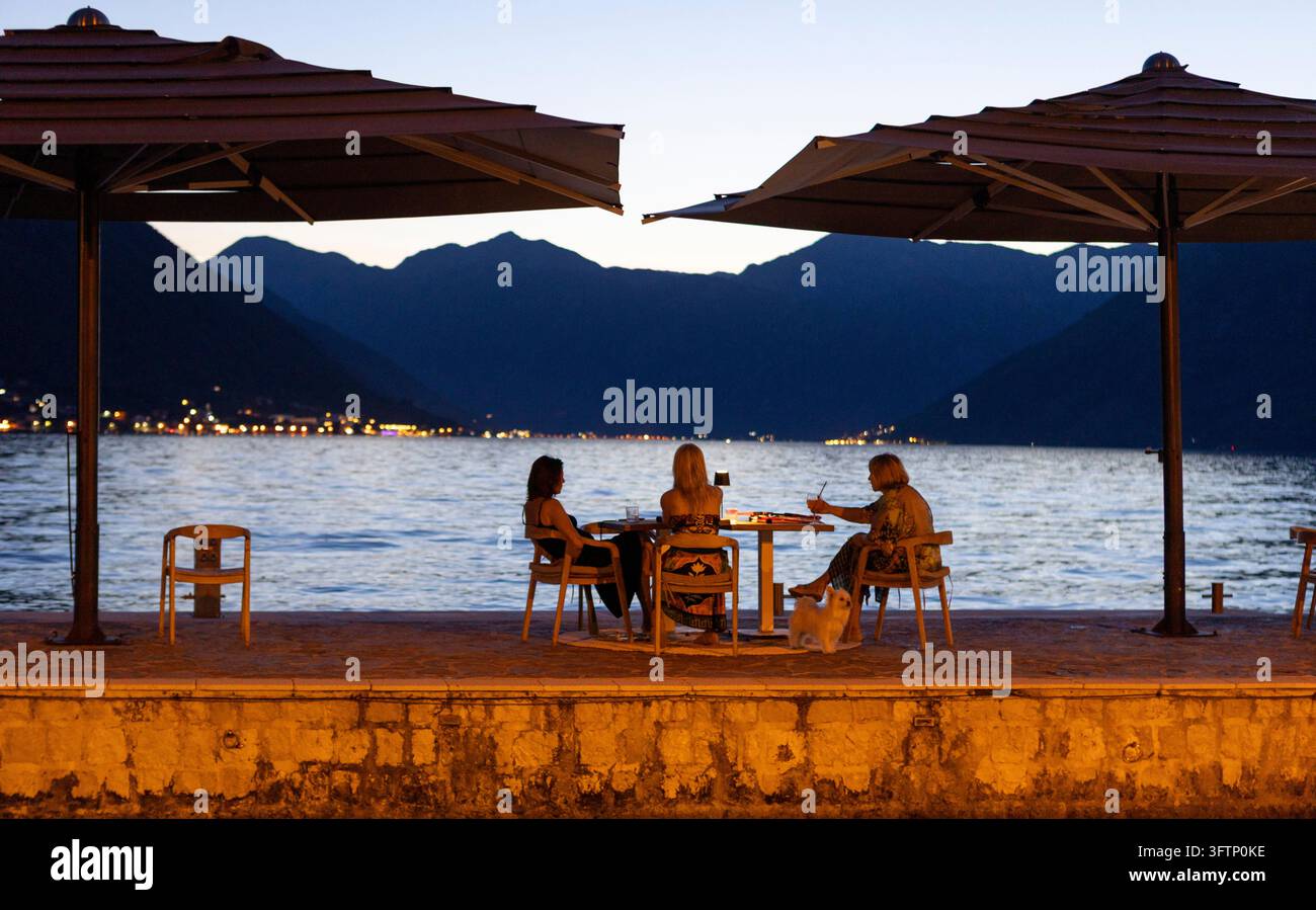 5 juillet 2026, Kotor, Monténégro, Trois femmes sont assises à une table au bord de l'eau, profitant d'une soirée paisible. Elles sont sous de grands parasols, avec des montagnes Banque D'Images