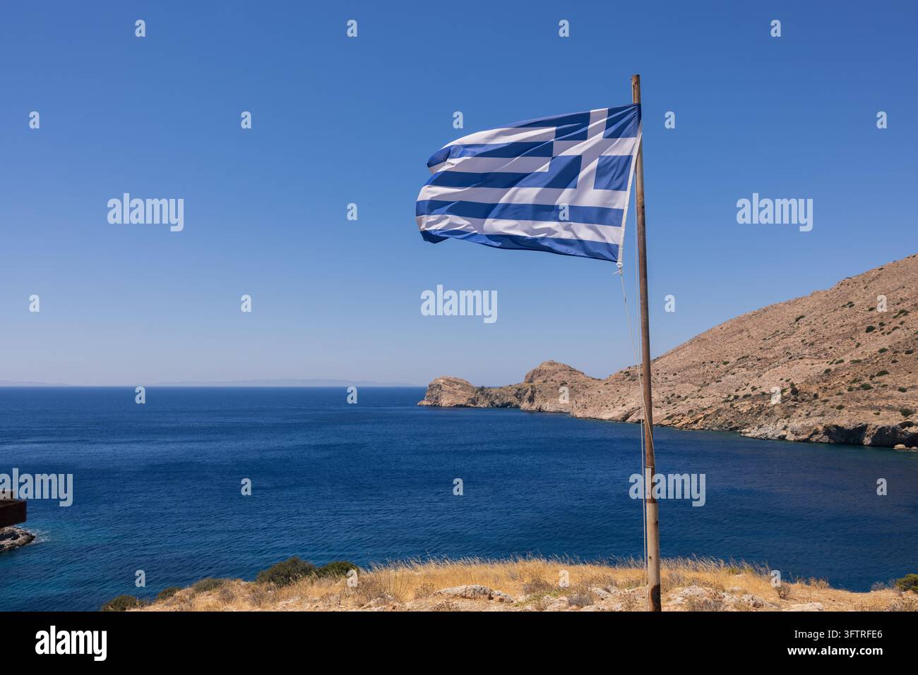 Le drapeau grec flotte au-dessus de la côte rocheuse près de la chapelle Agia Pakou sur l'île de Syros, surplombant les eaux bleu profond de la mer Égée et la rugg Banque D'Images
