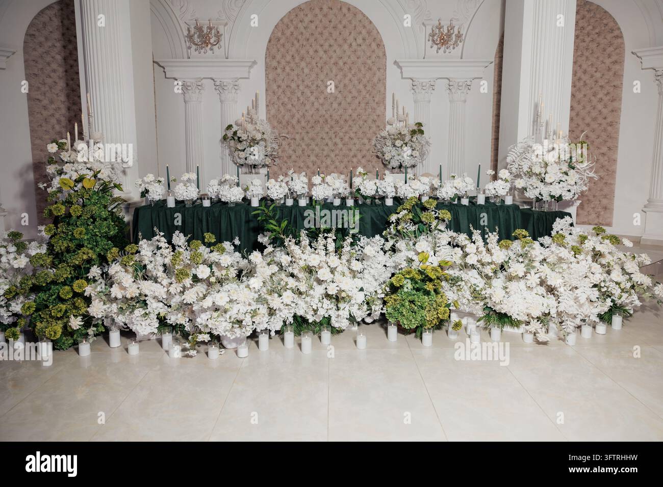 Table de banquet avec des arrangements floraux blancs luxuriants et du feuillage vert dans une salle de réception ornée, des bougies et des vases au sol et sur la table pour une élégance Banque D'Images