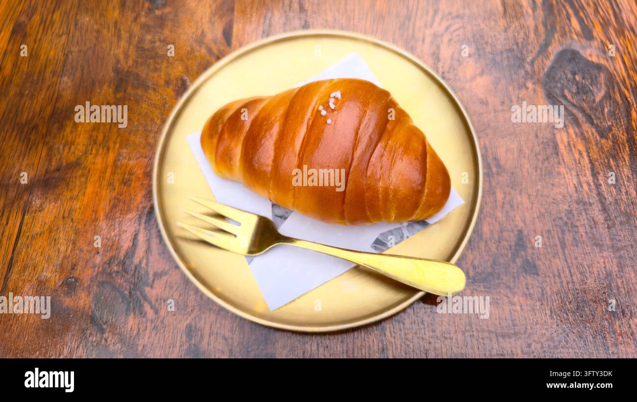 Pain salé japonais avec une croûte dorée brillante servi dans une assiette avec une fourchette sur une table en bois rustique, un pain de boulangerie moelleux et beurré populaire et en-cas de café - Image de stock capturée avec un smartphone