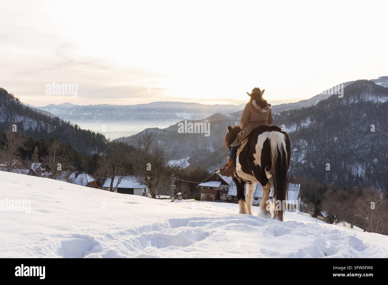 Vue arrière d'une femme montant un cheval à travers la neige en direction des montagnes, explorant la nature sauvage hivernale avec un esprit de nostalgie des voyages. Banque D'Images