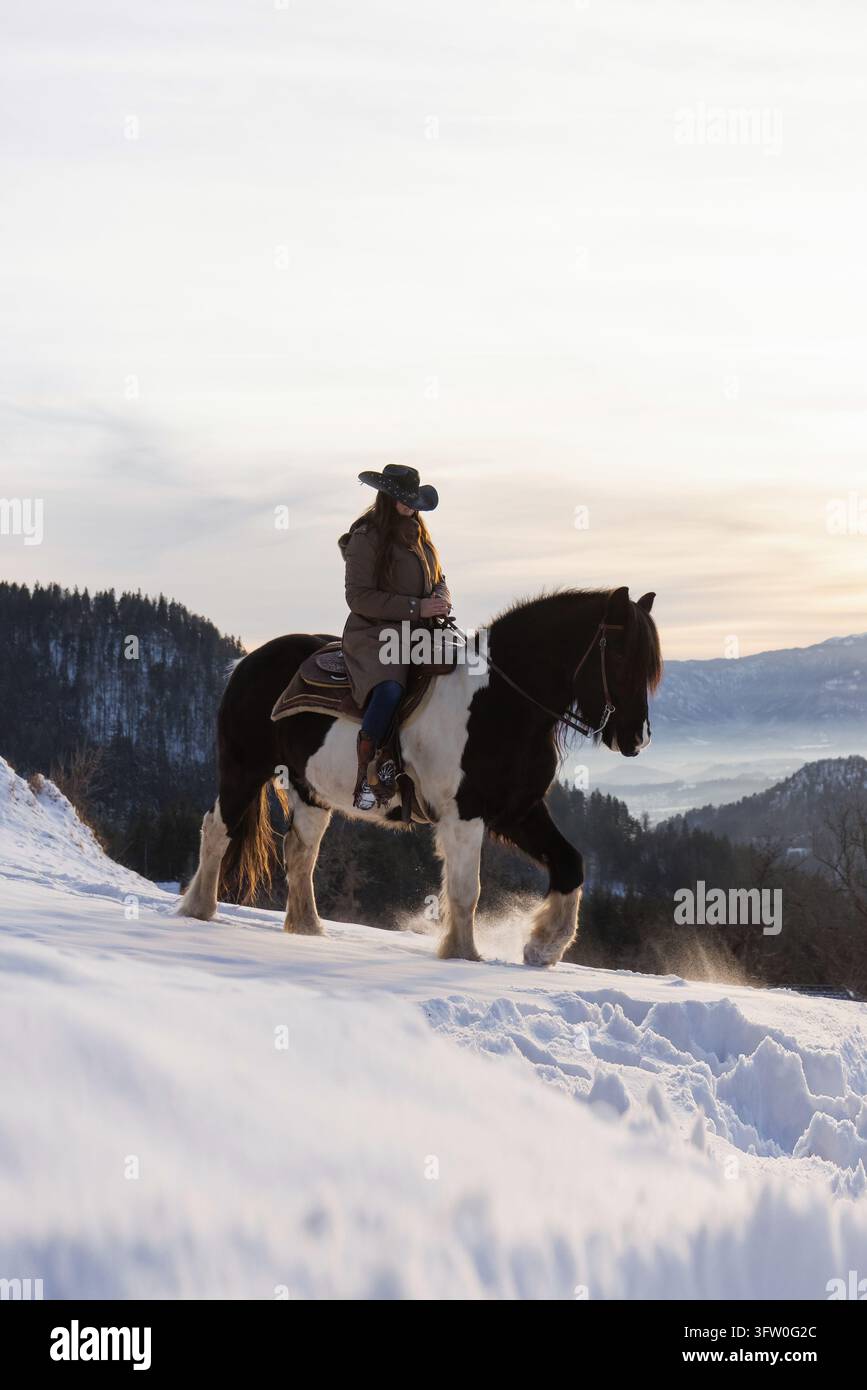 Femme montant à cheval dans la neige au coucher du soleil, entourée d'un paysage montagneux paisible et profitant d'un moment calme de connexion avec la nature. Banque D'Images