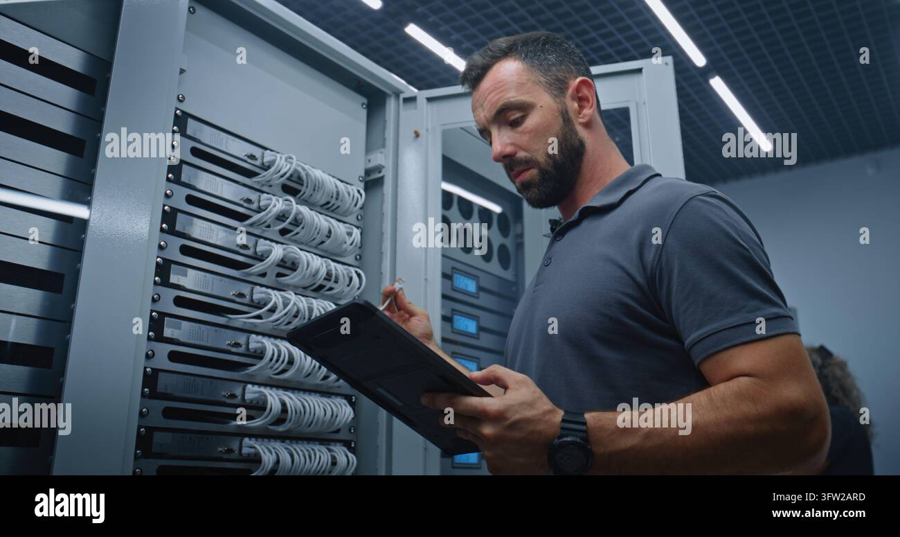 Spécialiste de salle serveurs masculin examinant les métriques des serveurs sur une tablette tout en inspectant les ports et connexions Ethernet, soutenant des performances réseau fiables, la maintenance des systèmes et les opérations numériques. Banque D'Images