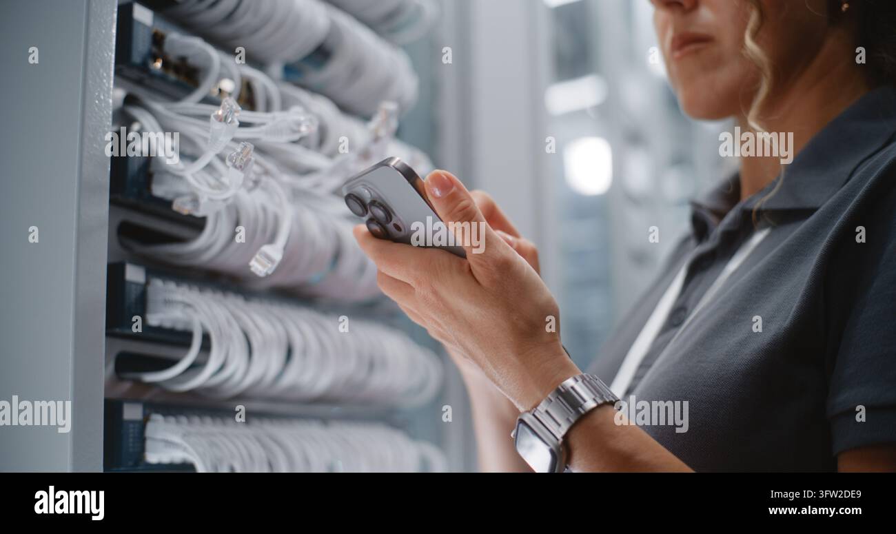 Technicienne de réseau féminine effectuant un audit physique des ports à côté d’un rack de serveurs, utilisant un smartphone pour vérifier l’identification des câbles et les enregistrements réseau. Concept de maintenance de l’infrastructure du centre de données. Banque D'Images