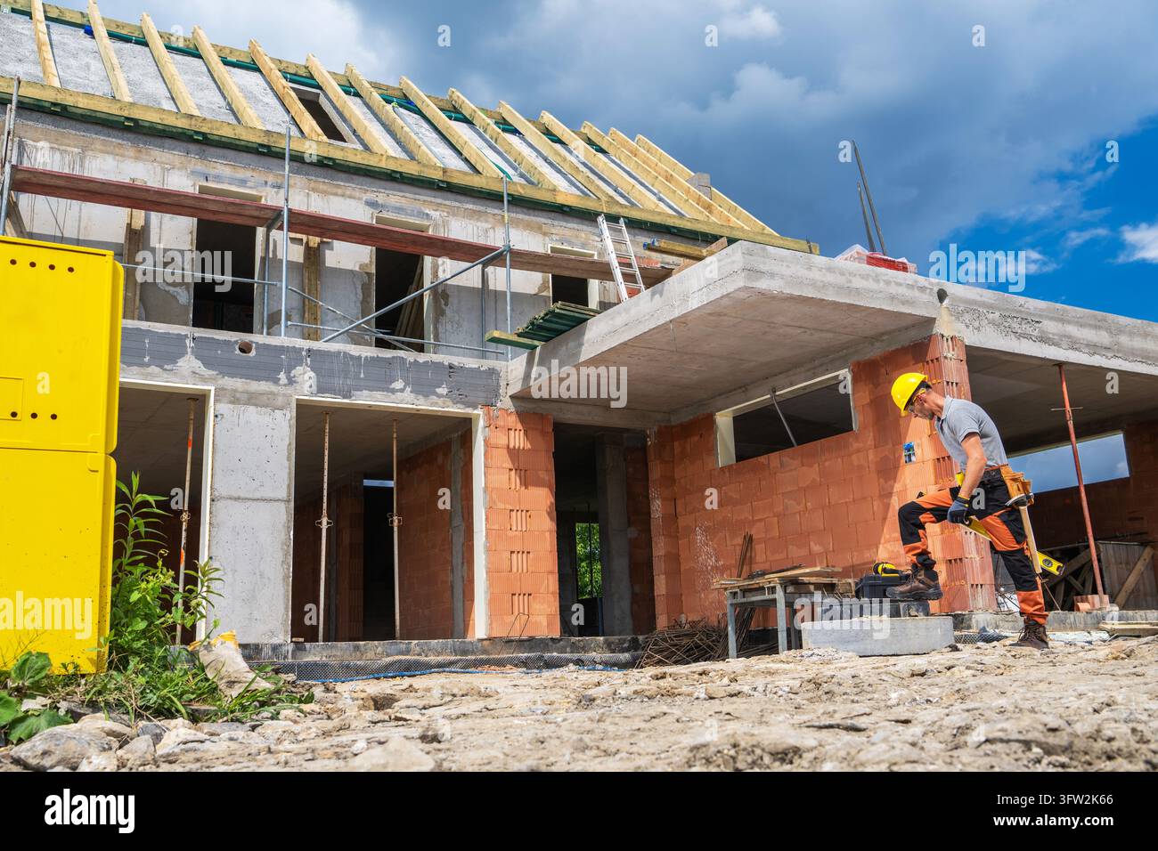 Un ouvrier portant un casque de protection se tient à côté d'une maison en briques inachevée avec des dalles en béton, un échafaudage et une charpente de toit en bois apparente. Banque D'Images