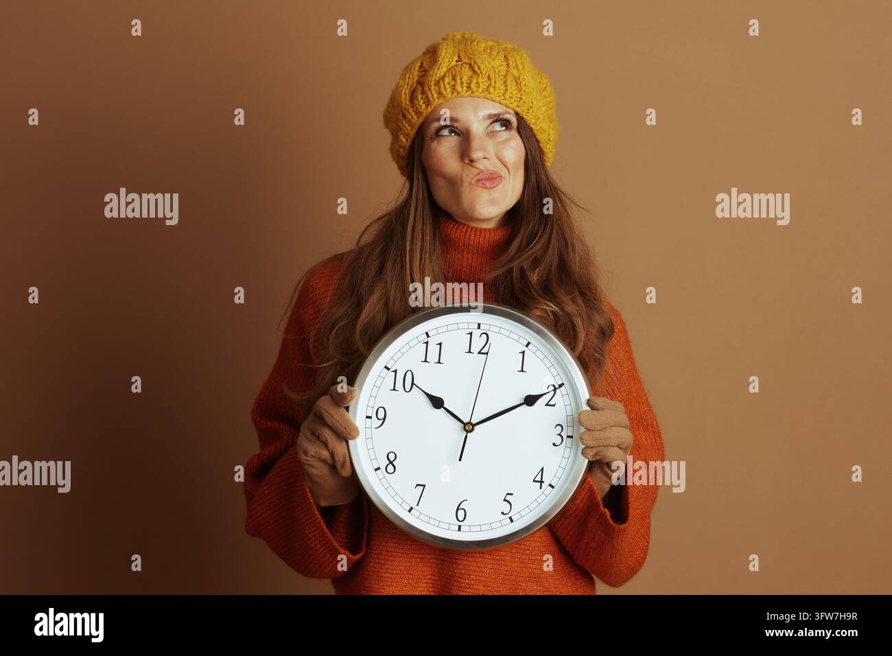 Une femme portant un bonnet jaune et un pull à col roulé orange tient une grande horloge murale, regardant vers le haut avec une expression pensive. La scène évoque la contemplation Banque D'Images