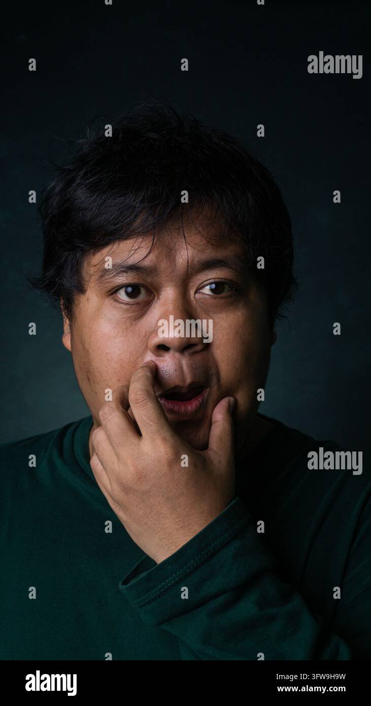 Un modèle masculin asiatique adulte affiche une expression faciale curieuse et légèrement confuse tout en reposant un doigt près de sa lèvre supérieure. Le sujet regarde di Banque D'Images