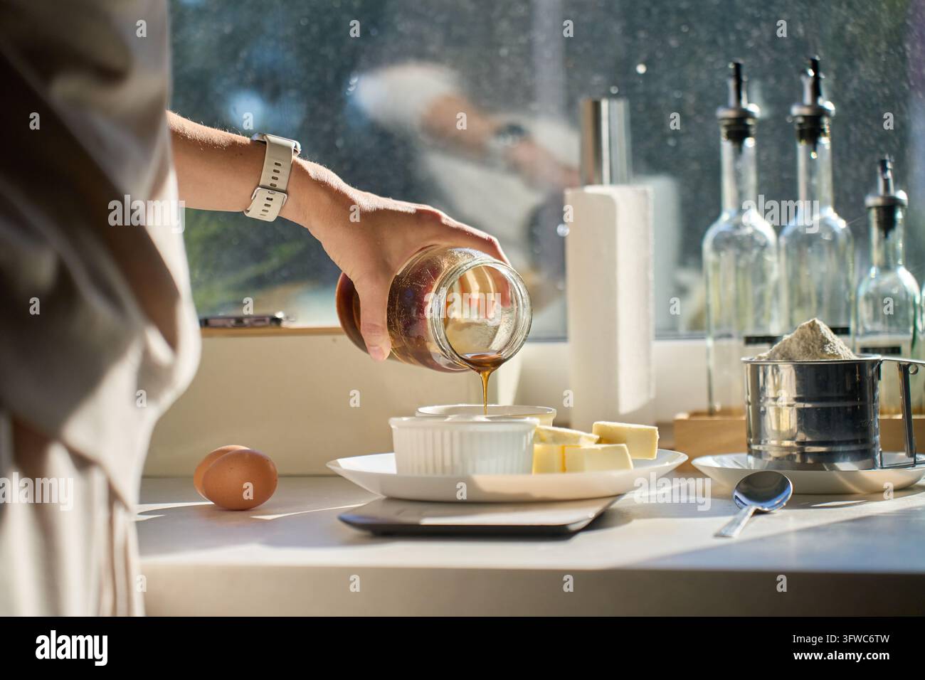 Femme versant du miel d'un bocal en verre dans un bol dans la cuisine. Beurre, œuf et farine sur la table pour faire un gâteau maison. Pâtisserie maison et préparation de desserts. Banque D'Images
