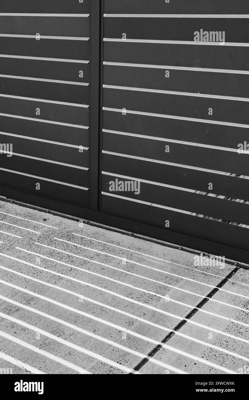 Abstrait noir et blanc de lattes de clôture métalliques horizontales projetant des rayures de lumière du soleil sur un pavé en béton Banque D'Images
