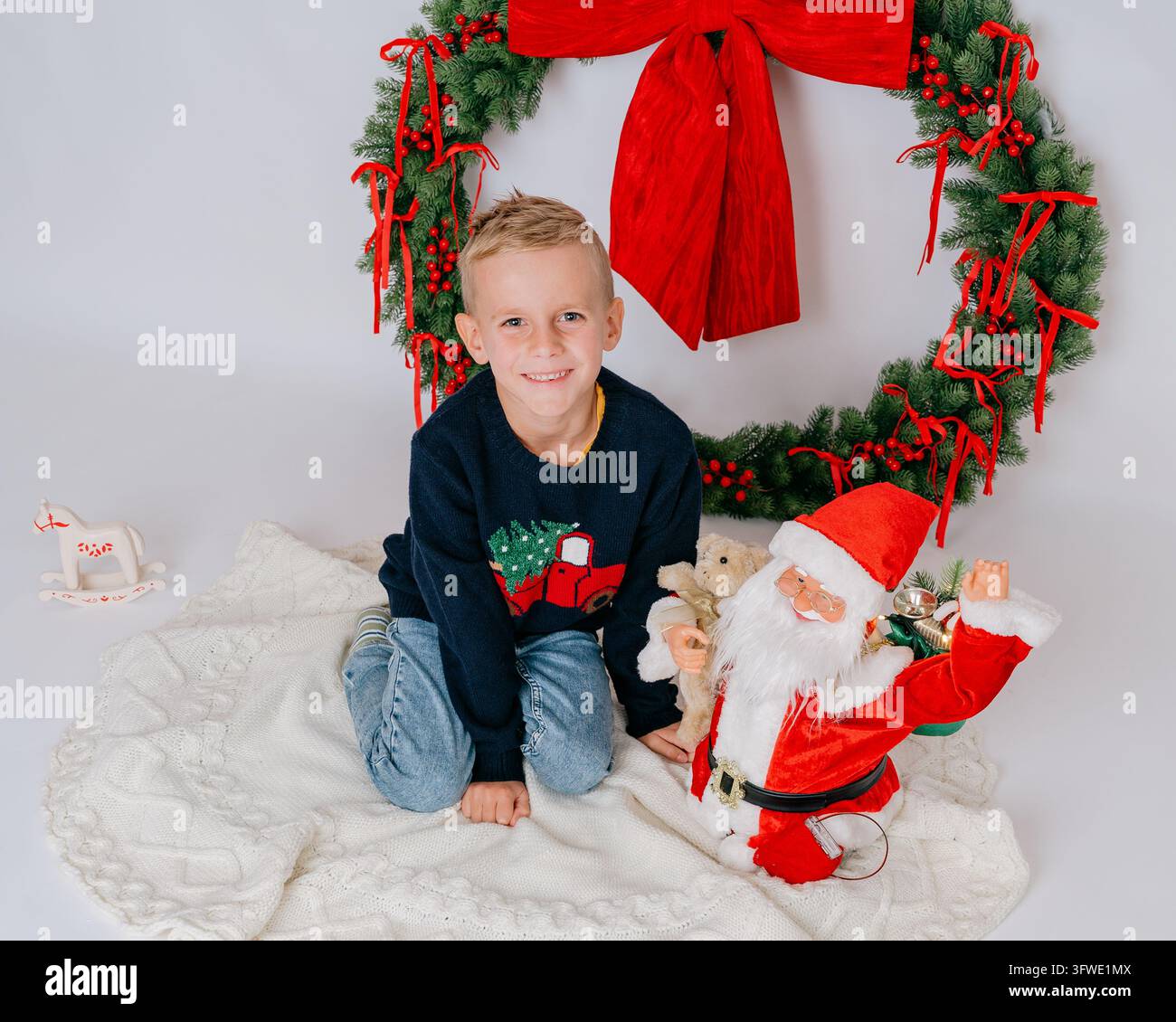 Garçon mignon en pull sombre assis sur une couverture blanche devant une grande couronne de Noël avec un nœud rouge et un jouet du Père Noël à côté de lui. Cadre de studio propre Banque D'Images