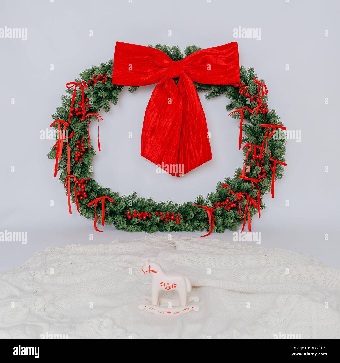 Grand couronne de Noël éternelle avec un grand nœud en velours rouge et des rubans attachés, posée sur fond blanc avec un petit cheval à bascule en bois. Motifs festifs minimalistes Banque D'Images