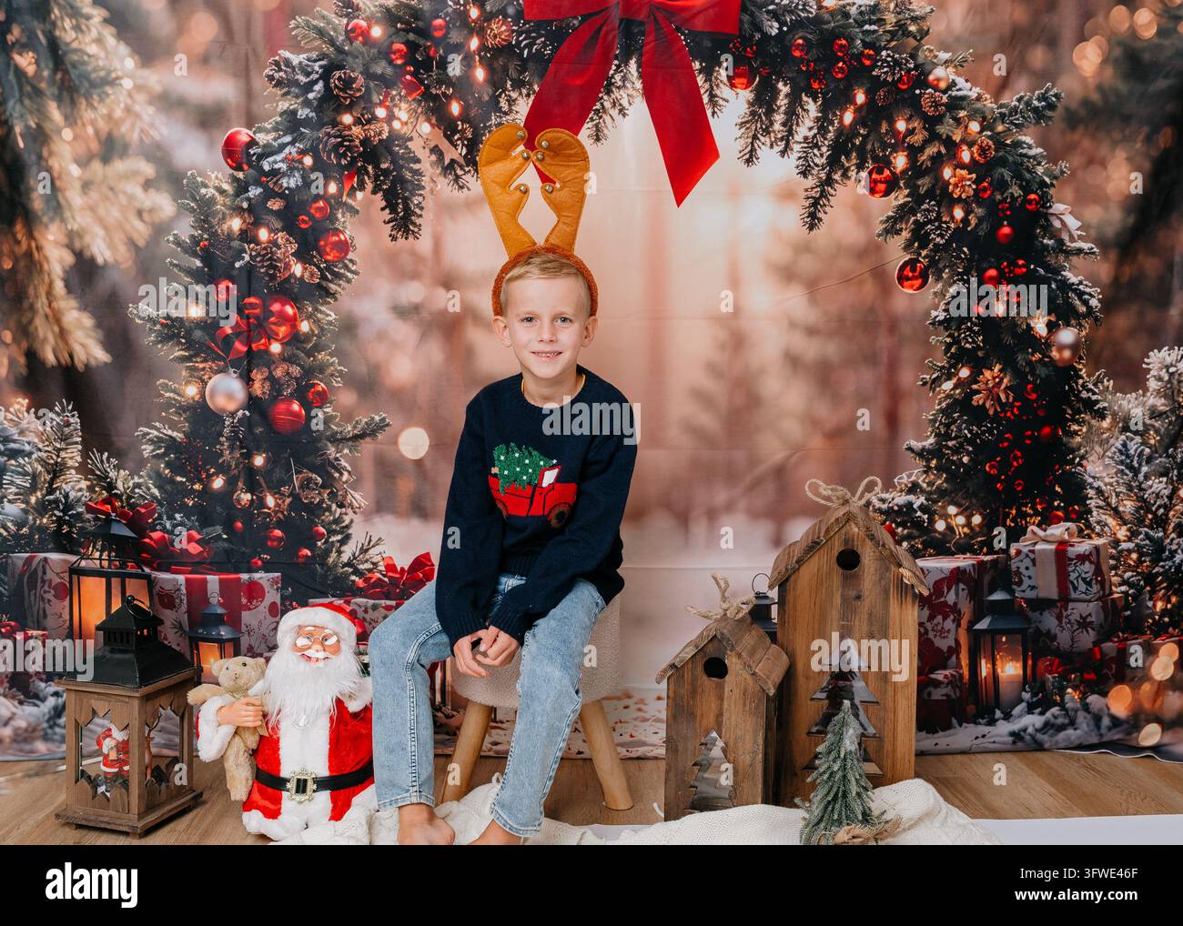 Portrait d'un garçon heureux portant un bandeau rouge à rennes assis sur un banc en bois dans un studio photo de Noël cosy. Fond d'hiver avec couronne d'épicéa Banque D'Images