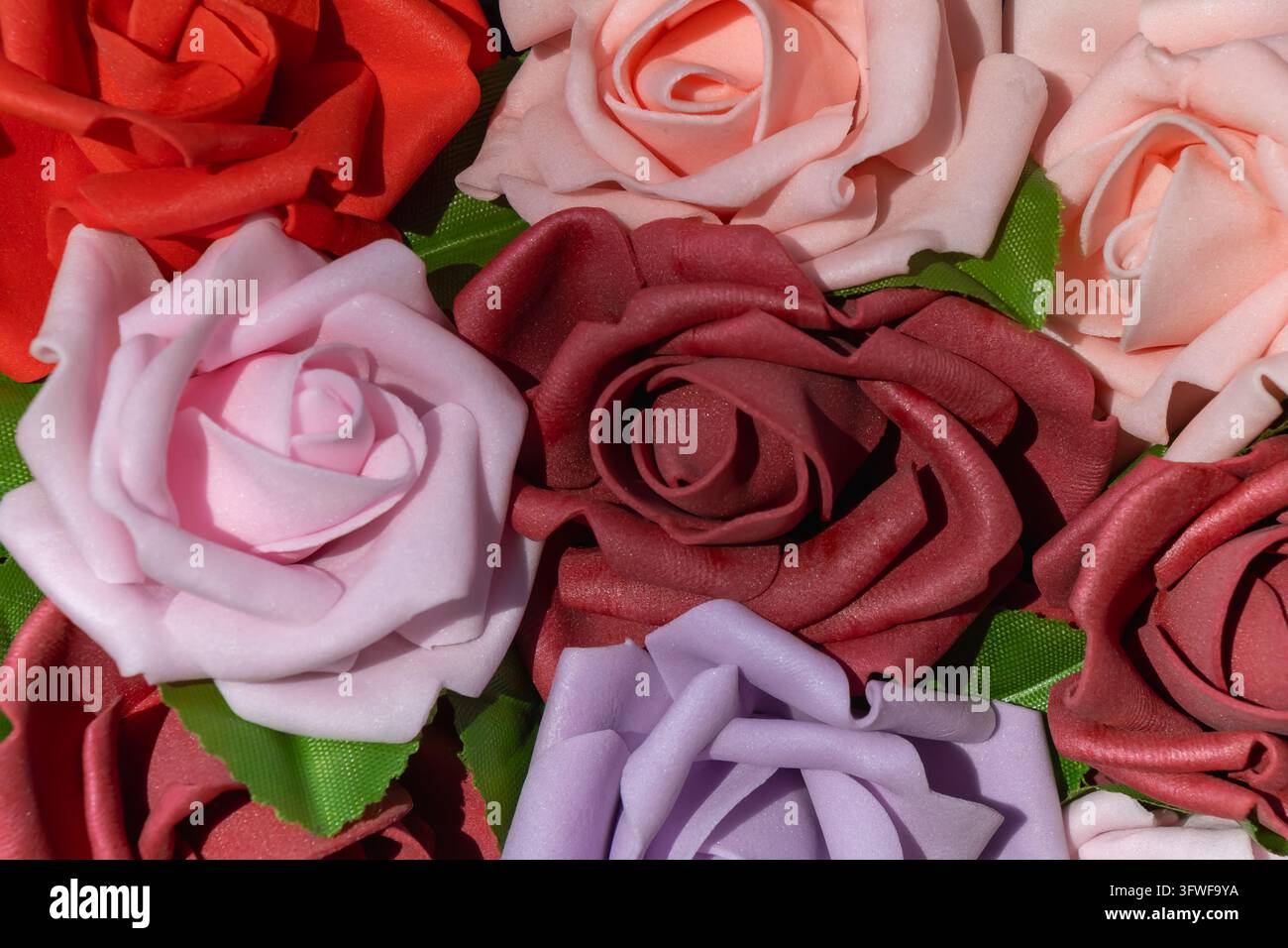 Close-up de roses artificielles en rouge, rose, bordeaux et lilas avec des feuilles vertes, créant un arrangement floral coloré et dense et un objet artisanal décoratif Banque D'Images
