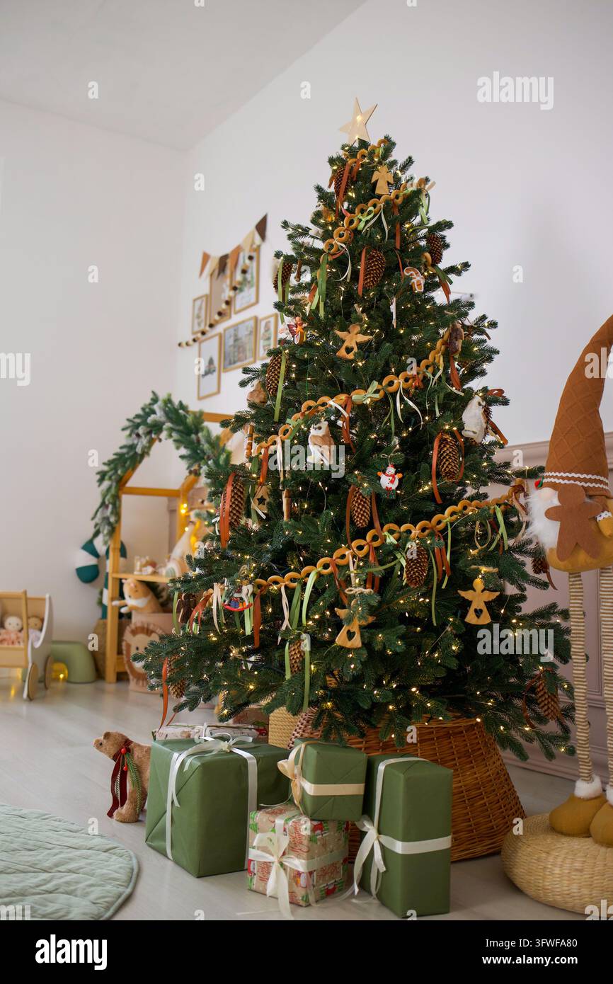 Arbre de Noël décoré avec des guirlandes en bois, des ornements en pommes de pin, des guirlandes lumineuses chaudes et des cadeaux emballés dans une chambre d'enfant confortable Banque D'Images