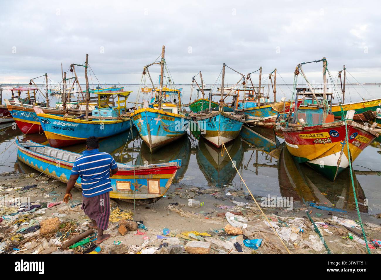 Pêcheur marchant vers des bateaux de pêche colorés sur une rive jonchée de déchets, Province du Nord, Jaffna, Sri Lanka Banque D'Images