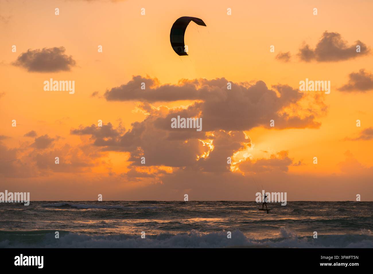 Vue d'un kitesurfeur sur une planche à hydrofoil sur l'océan pendant un coucher de soleil doré vibrant avec des nuages dans le ciel, Mexique. Banque D'Images