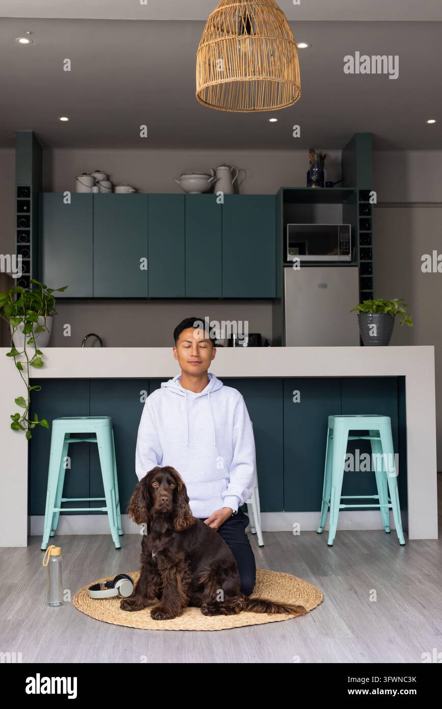 Homme asiatique d'âge moyen portant un sweat à capuche et un pantalon, agenouillé, caressant un chien brun sur un tapis tissé dans la cuisine Banque D'Images