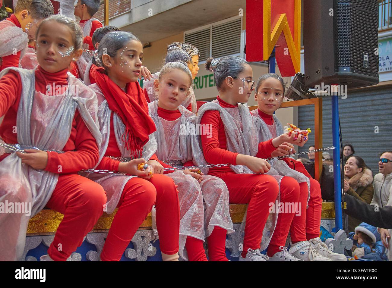 Cinq filles en tenues rouges et tuniques argentées translucides sont assises en rangée sur un char décoré, tenant des bonbons et regardant dans différentes directions Banque D'Images