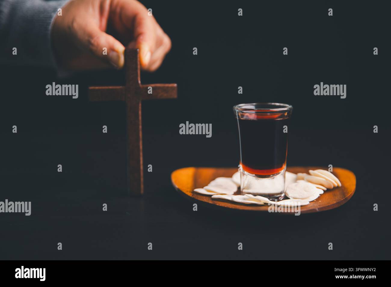 Main tenant une croix en bois à côté d'hosties et d'une boisson rouge foncé sur une assiette en bois, représentant le culte chrétien, la foi, la prière, le souvenir sacré Banque D'Images
