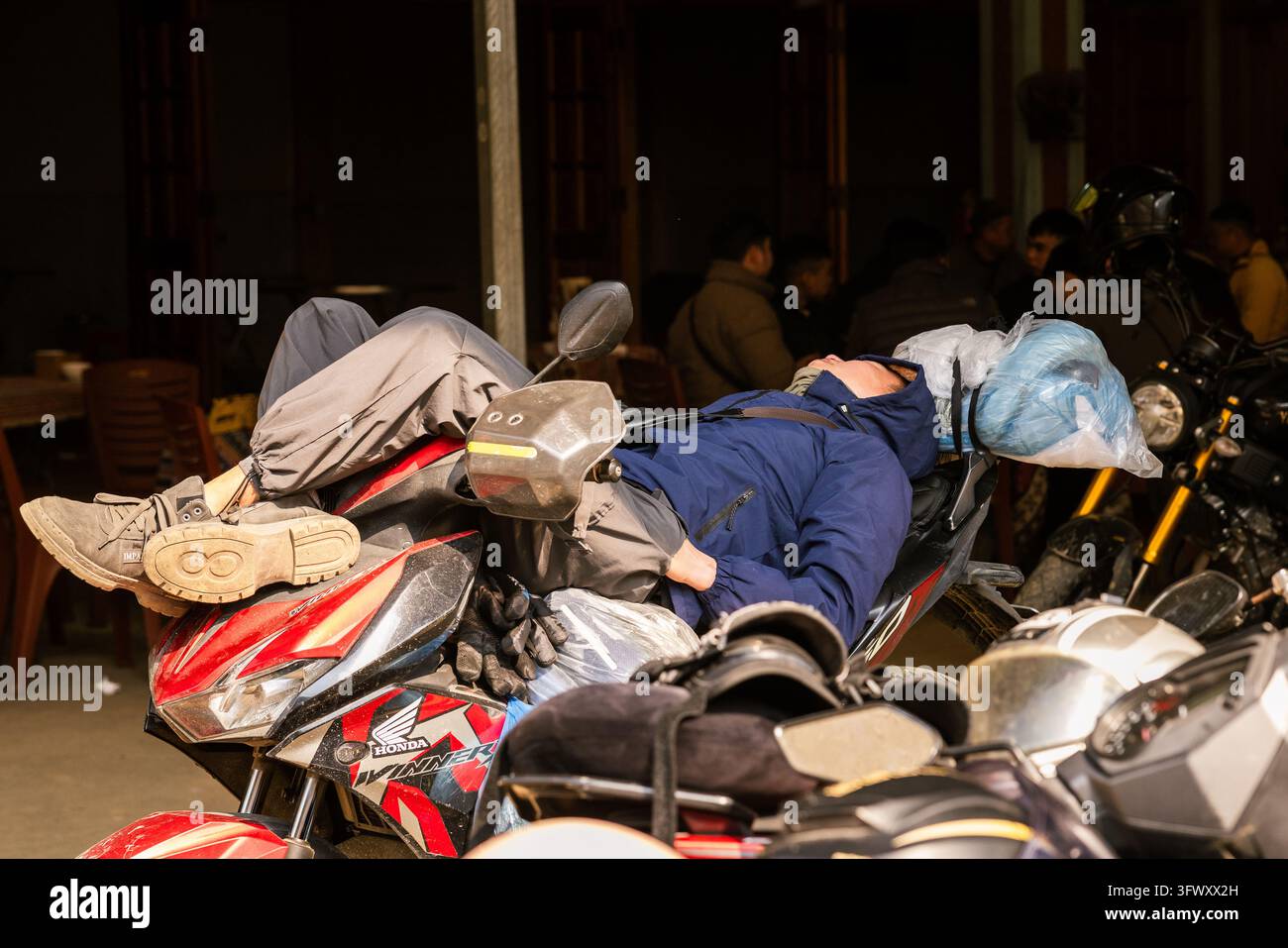 Ha Giang, Vietnam - 12 janvier 2026 : Un homme en veste bleue est allongé et se repose sur une moto Honda rouge dans une zone faiblement éclairée avec d'autres... Banque D'Images