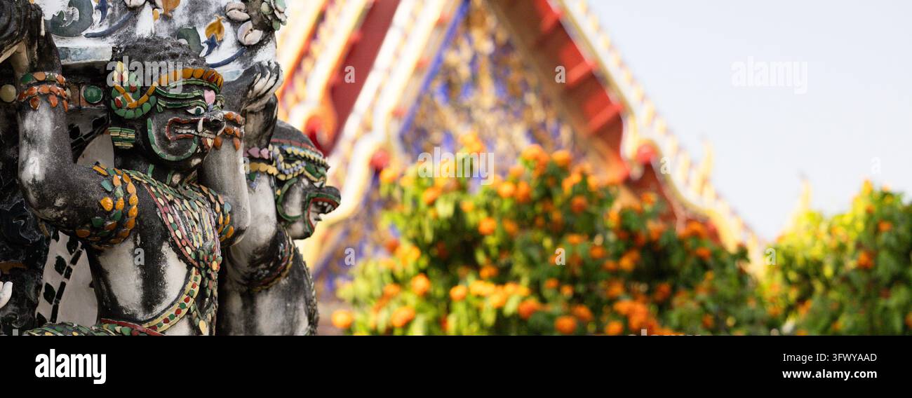Bangkok, Thaïlande - 1er février 2026 : Des statues de gardiens colorées se dressent devant un toit de temple flou et des orangers. Banque D'Images
