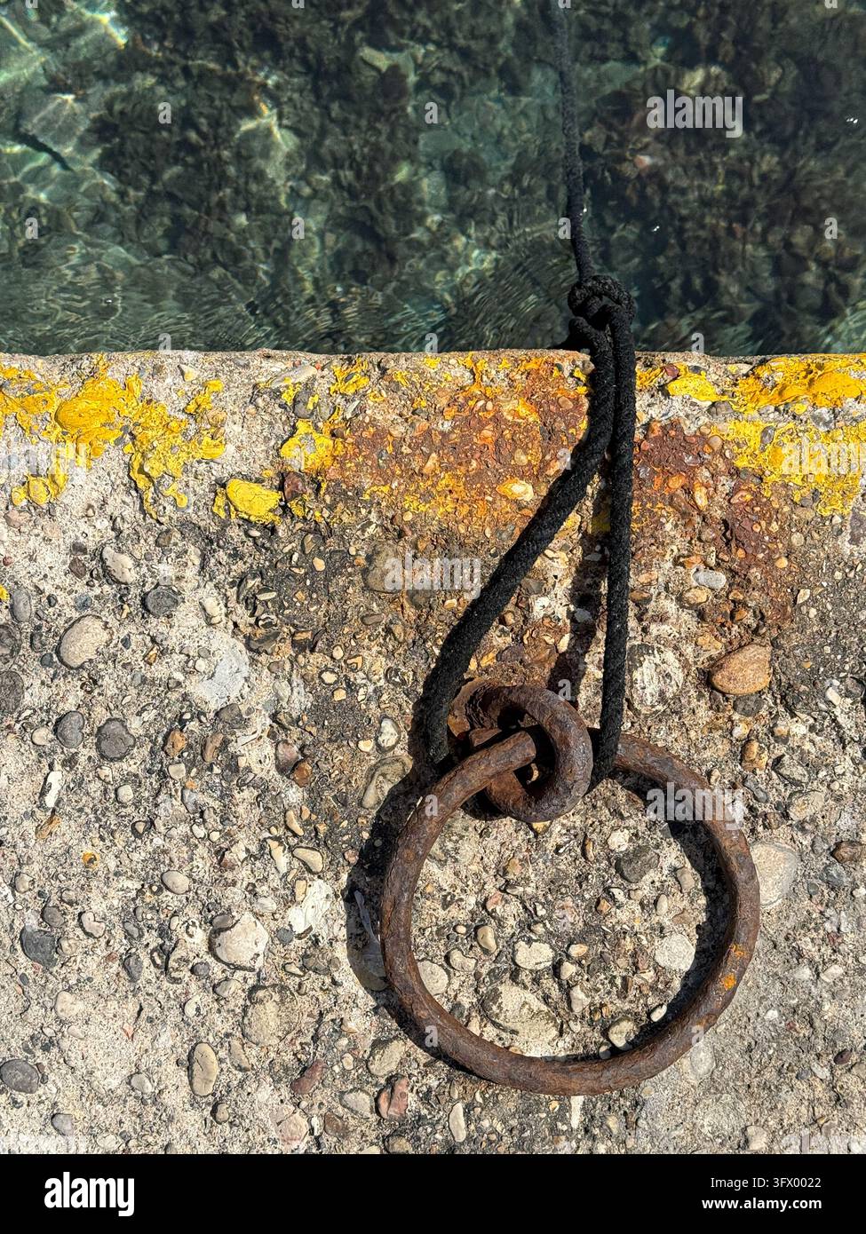 Une corde attachée à un anneau d'amarrage rouillé repose sur un quai en pierre à côté d'une eau claire à Logos, Îles Ioniennes, Grèce, pendant la journée. Banque D'Images
