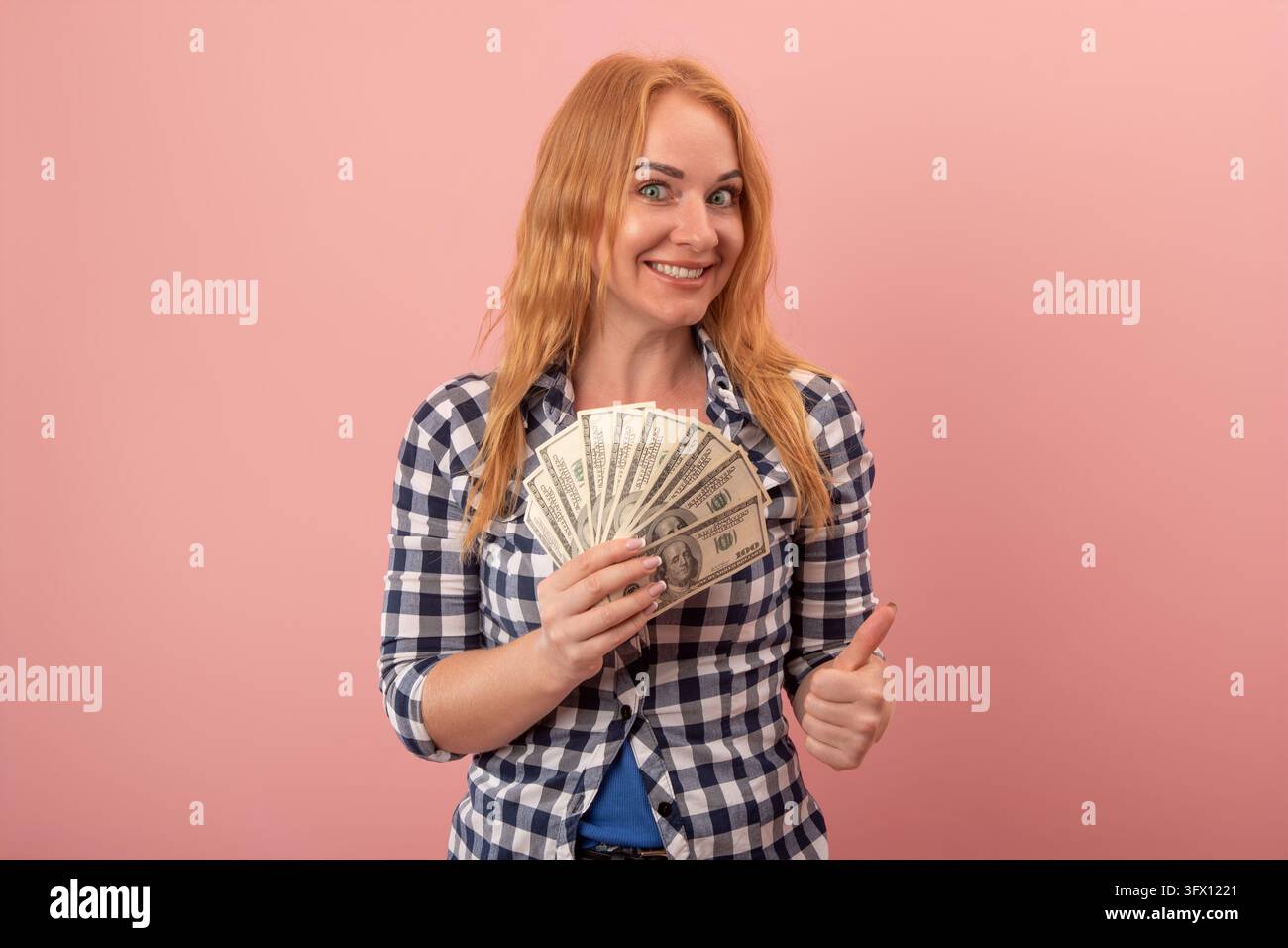 Jeune femme joyeuse aux cheveux roux exprimant le succès et l'approbation, tenant un éventail de billets de banque des États-Unis et montrant un geste positif sur un Banque D'Images