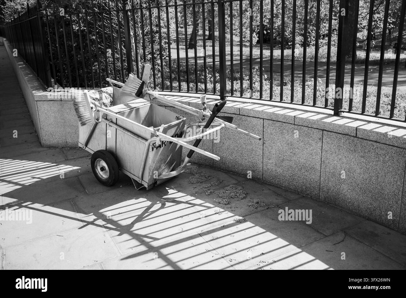 CHARIOT DE NETTOYAGE DE RUE À CÔTÉ DES RAILLES DU PARC EN NOIR ET BLANC, LONDON HYDE PARK BAYSWATER RÉPORTAGE DE VOYAGE, LONDON, ANGLETERRE : Un chariot de maintenance vide équipé de brosses et d'une pelle est posé sur une allée pavée à côté de rampes en fer projetant des ombres nettes. INFO : La zone de Hyde Park et Bayswater combine de vastes espaces verts avec un charme cosmopolite dans l'ouest de Londres. Le magnifique Hyde Park offre des promenades en bateau et des sentiers arborés, tandis que le voisin Bayswater présente une architecture victorienne majestueuse. Banque D'Images