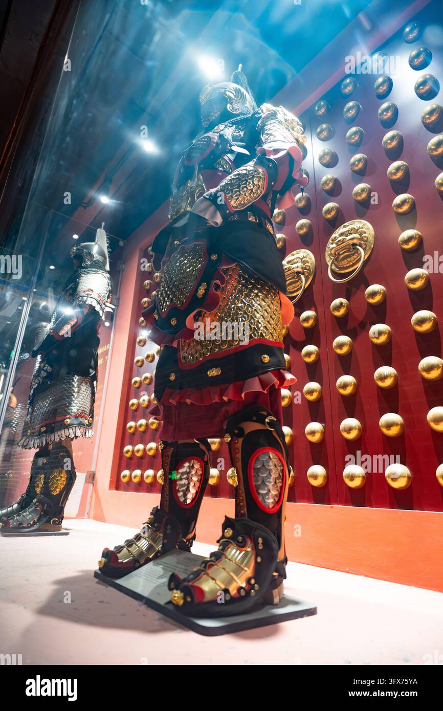 Armure traditionnelle japonaise de samouraï exposée dans une vitrine muséale, comprenant des plaques lamellaires laquées ornées, une jupe kusazuri complexe, des protections de cuisses haidate, des gardes de tibias suneate, et un casque kabuto décoratif avec crête maedate, placé devant une porte laquée rouge avec des bossages de style tsuba dorés et des poignées en bronze à anneaux, illuminé par des projecteurs dramatiques créant des reflets de lumière et une brume atmosphérique Banque D'Images