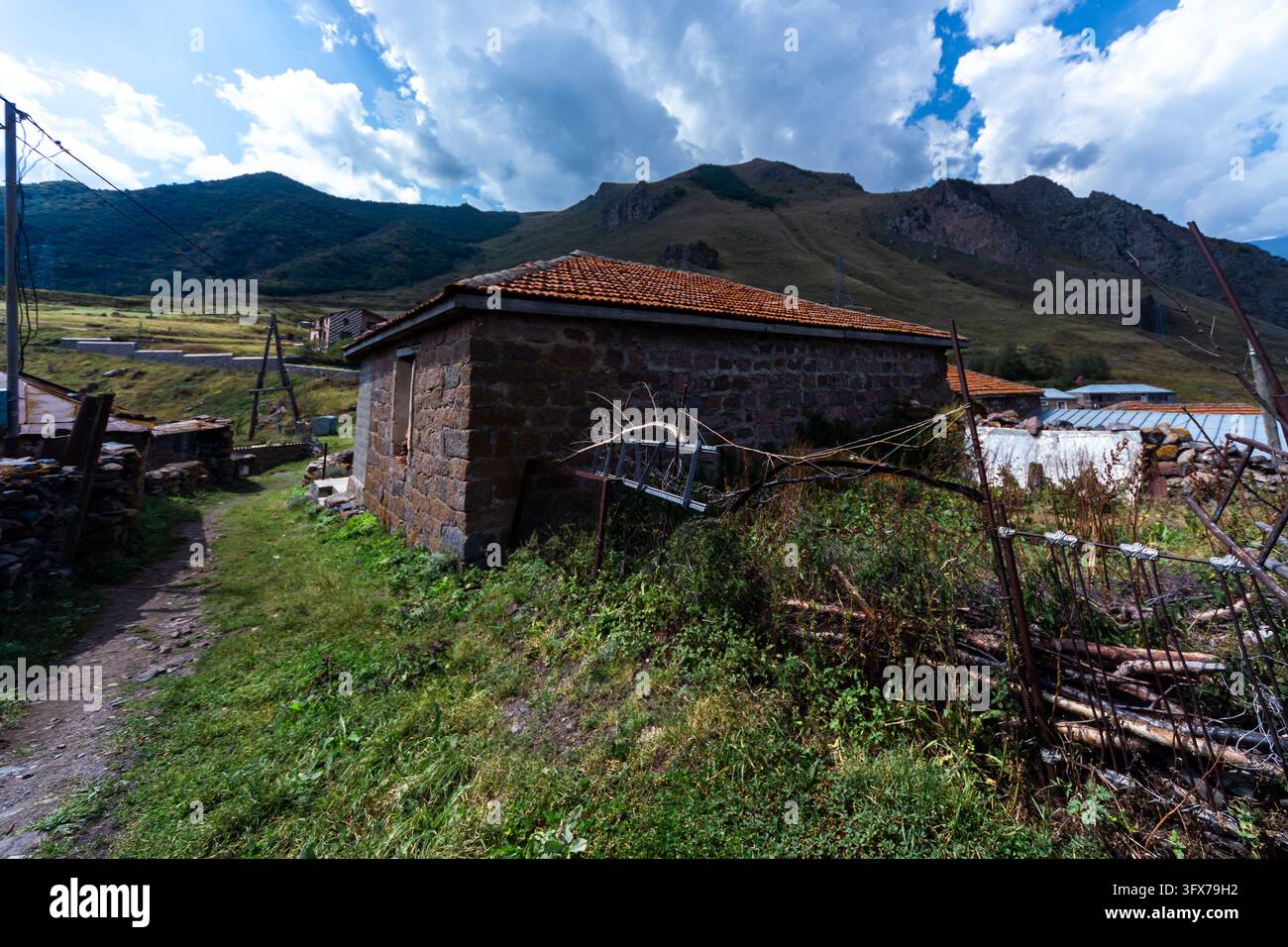 Une maison rustique historique construite en blocs de pierre naturelle présente un toit en tuiles orange niché parmi l'herbe verte et les montagnes sous un ciel nuageux. Banque D'Images