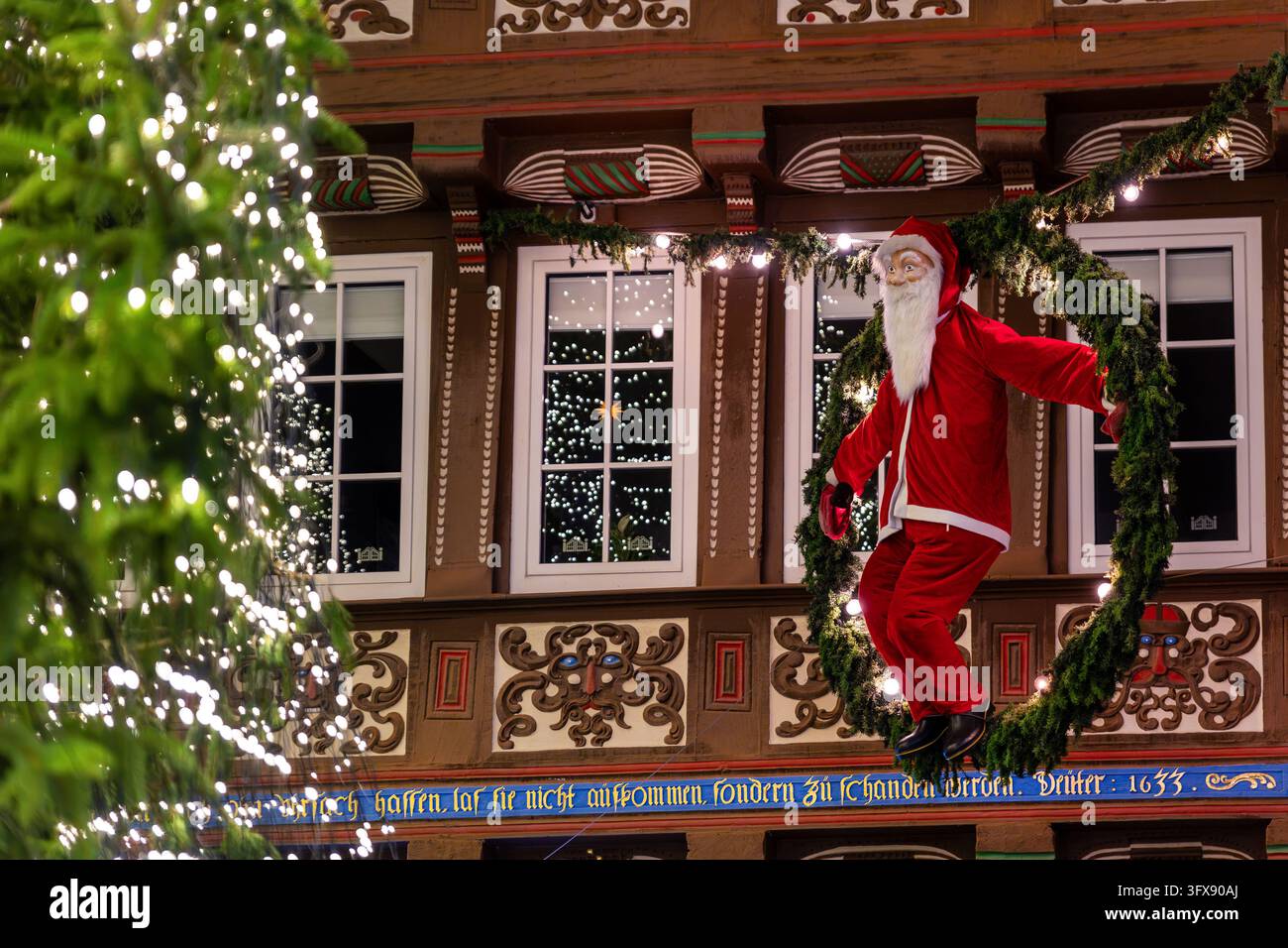 Figure du Père Noël en costume rouge suspendue à côté d'un sapin de Noël illuminé devant une façade en bois historique ornée. Guirlandes festives et fenêtres lumineuses Banque D'Images