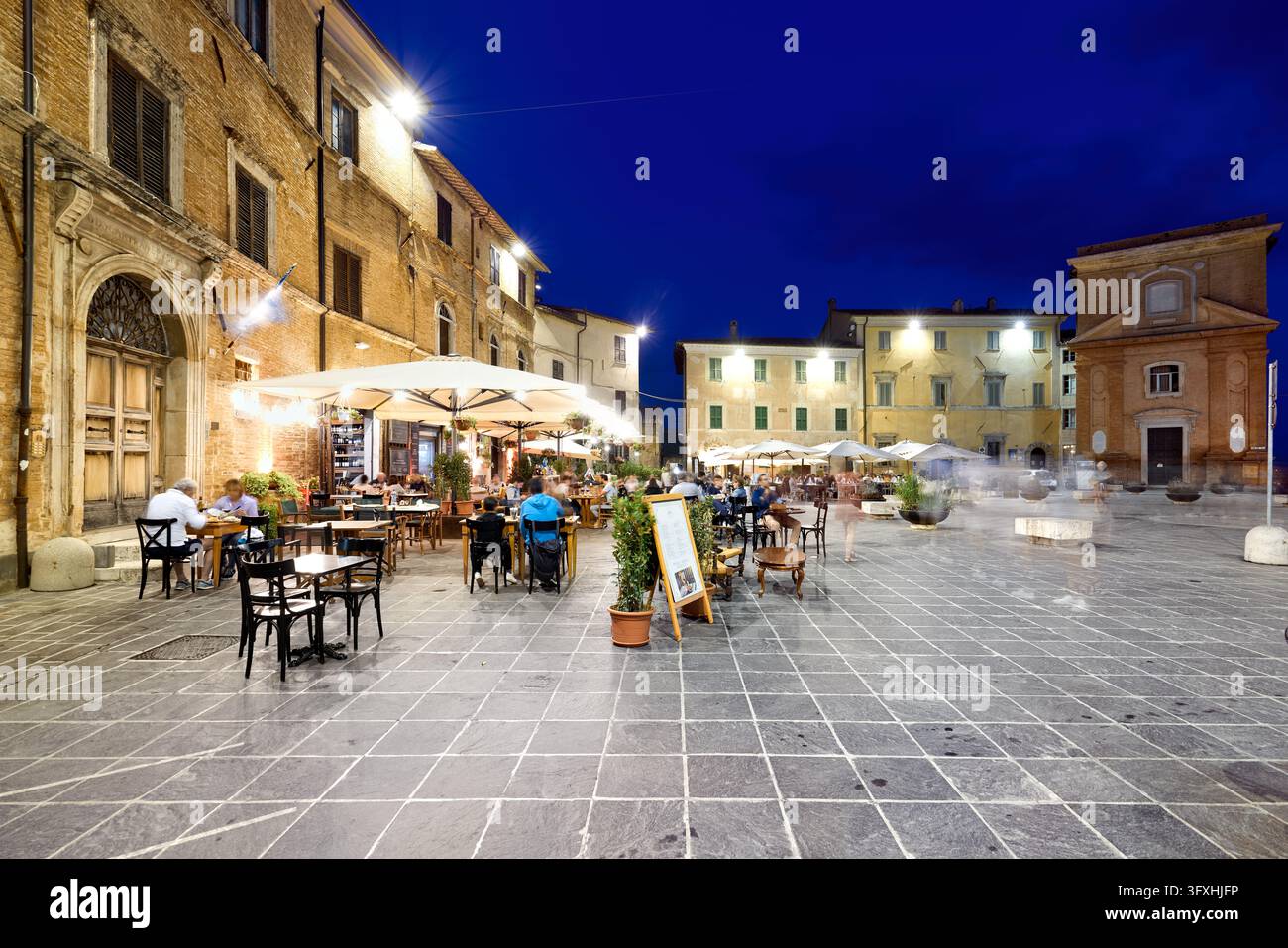 Les clients sont assis à des tables de restaurant en plein air sous des parasols blancs dans une place historique en briques en Ombrie, en Italie, pendant l'heure bleue au crépuscule. Banque D'Images