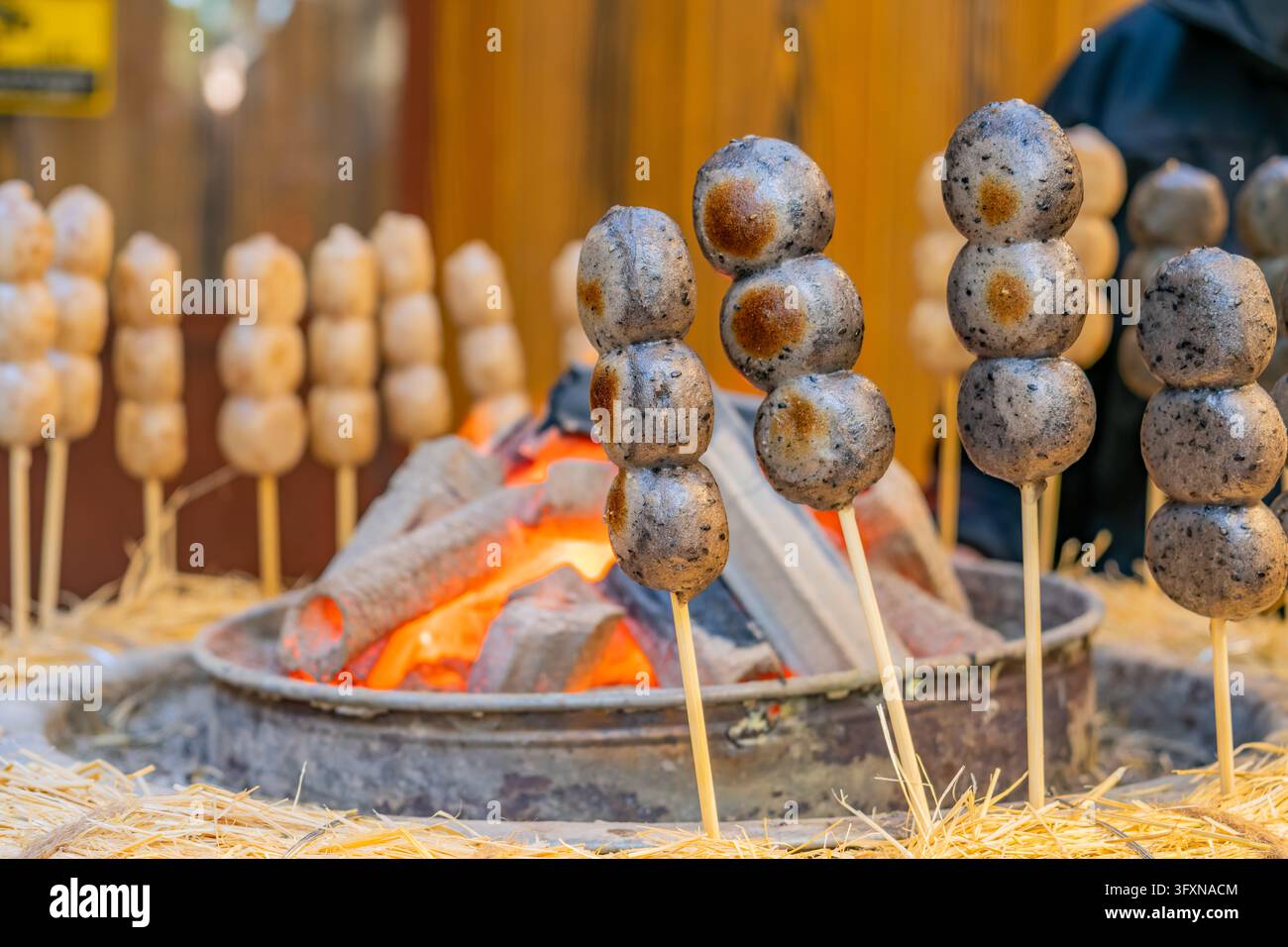 Tokyo, Japon, 22 NOV 2024 - Des brochettes de boules de dango sombres et tachetées sont disposées verticalement dans de la paille près d'un brasero en métal contenant du charbon ardent. Banque D'Images