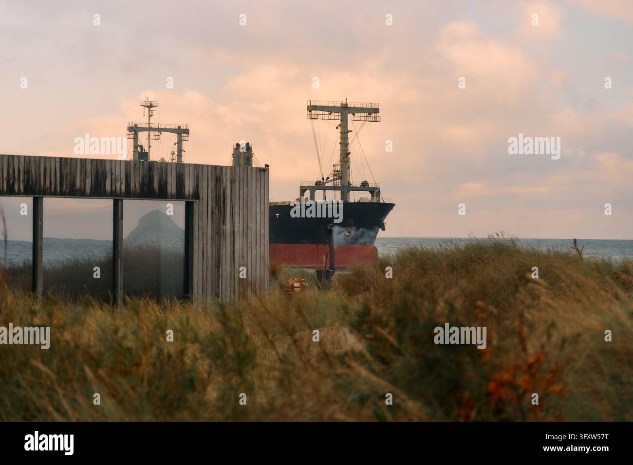 Navire de charge ancré au large près d'un bâtiment de plage en bois patiné encadré par des prairies de dunes au coucher du soleil, ciel pastel et scène côtière sombre, maritime Banque D'Images