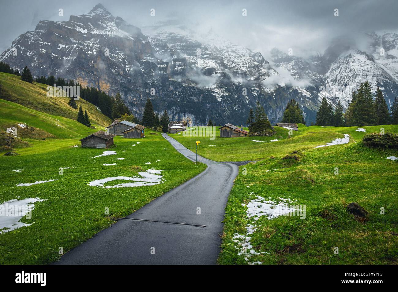 Route étroite et sinueuse sur le pâturage alpin avec de jolies cabanes en bois et des montagnes brumeuses spectaculaires en arrière-plan, Murren, vallée de Lauterbrunnen, Suisse Banque D'Images