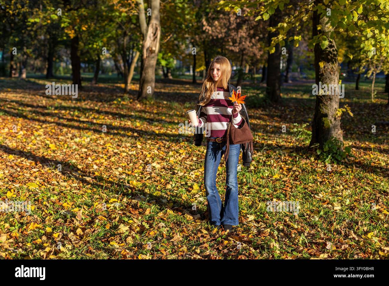 Jeune femme marchant dans un parc ensoleillé d'automne tenant une tasse de café à emporter et une feuille d'érable orange, profitant d'une journée paisible d'automne parmi les couleurs vives Banque D'Images