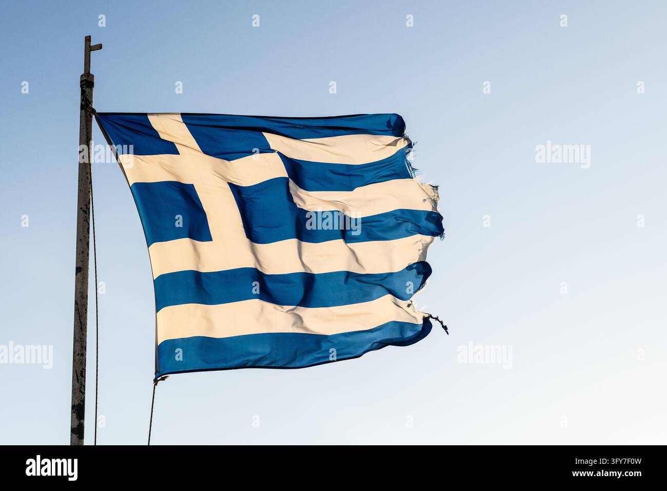 Un drapeau national grec usé, bleu et blanc aux bords effilochés, flotte fièrement dans le vent contre un ciel bleu clair, symbolisant la fierté nationale et Banque D'Images