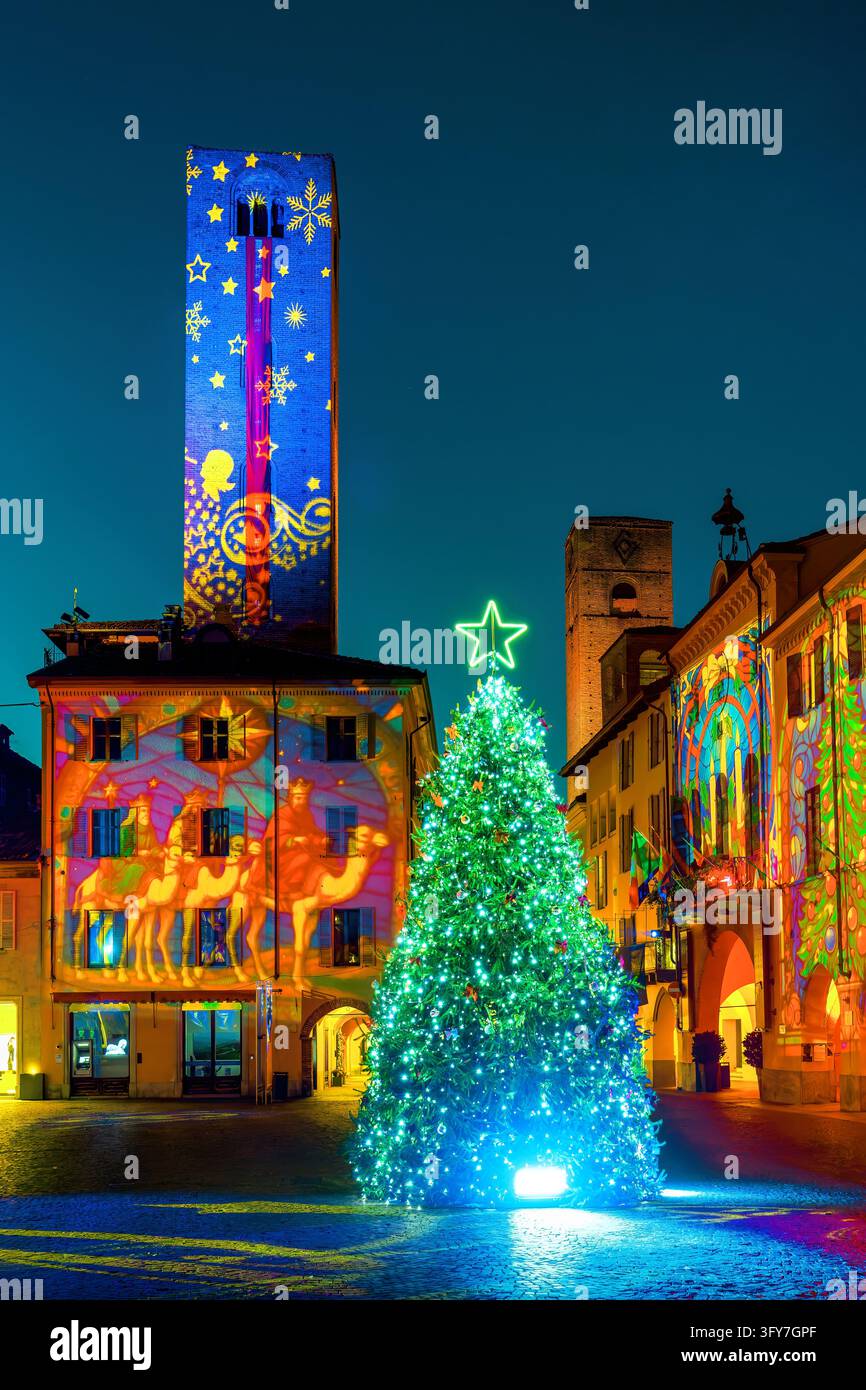 Sapin de Noël lumineux sur la place centrale avec des projections de lumières festives colorées sur la tour en briques médiévale et les façades historiques la nuit à Alba, en Italie Banque D'Images