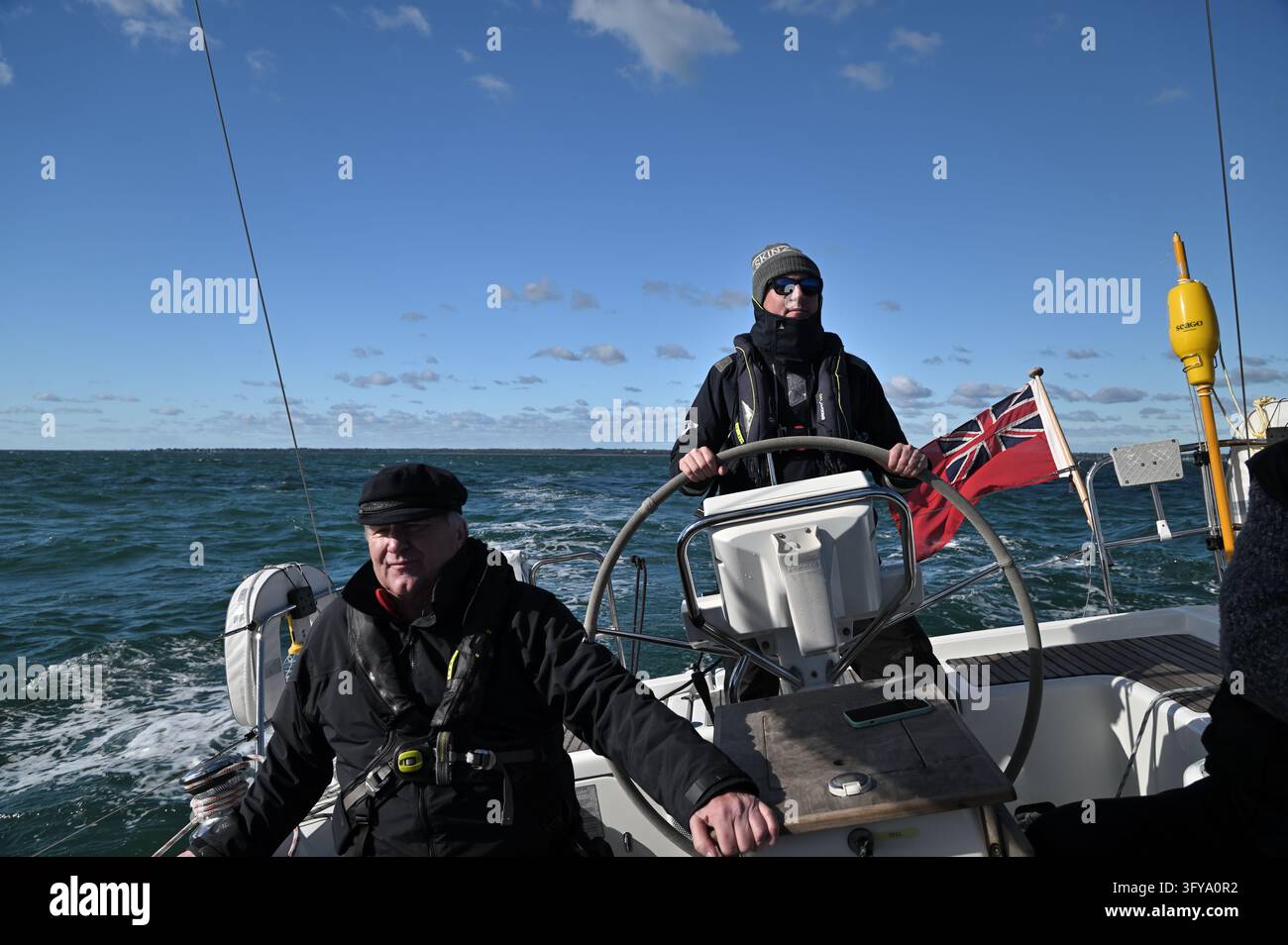 Marins au gouvernail d'un yacht arborant une enseigne rouge par une journée lumineuse et venteuse sur le Solent, Hampshire, Royaume-Uni, 17 octobre 2026 Banque D'Images