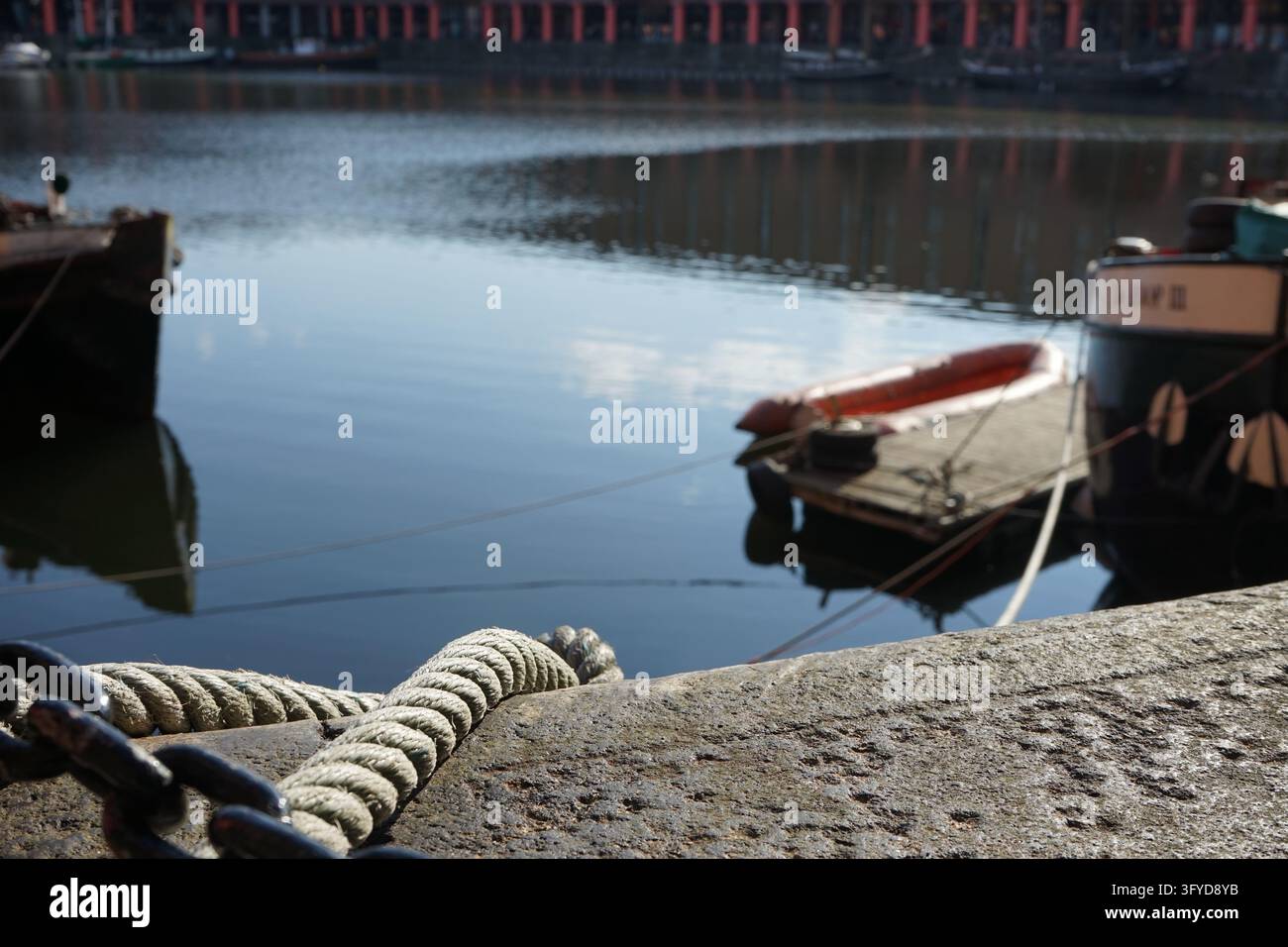 Une épaisse corde et une chaîne gisent sur le bord d'un quai en pierre avec des bateaux amarrés et une eau immobile en arrière-plan. Banque D'Images