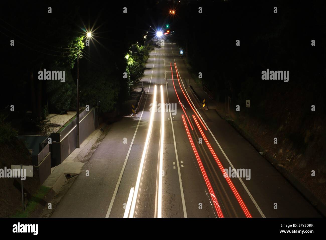 Traînées de lumière à exposition longue des feux des voitures blanches et rouges sur une route la nuit avec des lampadaires et des arbres sombres. Convient pour la circulation, le transport, la vie urbaine Banque D'Images