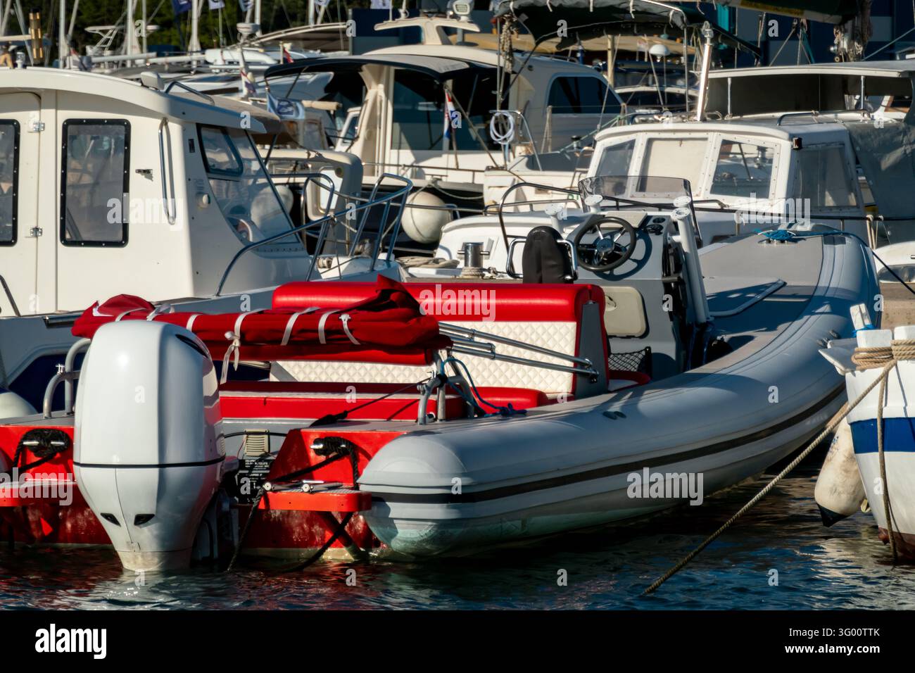 Bateau à moteur gonflable rouge et gris amarré parmi de petits croiseurs à cabine dans une marina animée, avec des moteurs hors-bord, des cordes et une eau calme. Banque D'Images