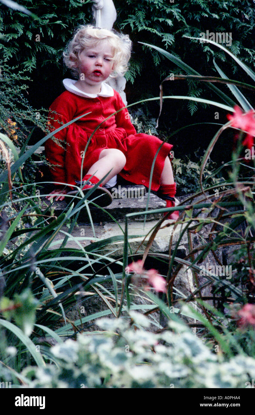 Petite fille aux cheveux blond vêtue de rouge, penchée contre UNE statue de jardin tout en étant assise près D'un étang. Banque D'Images