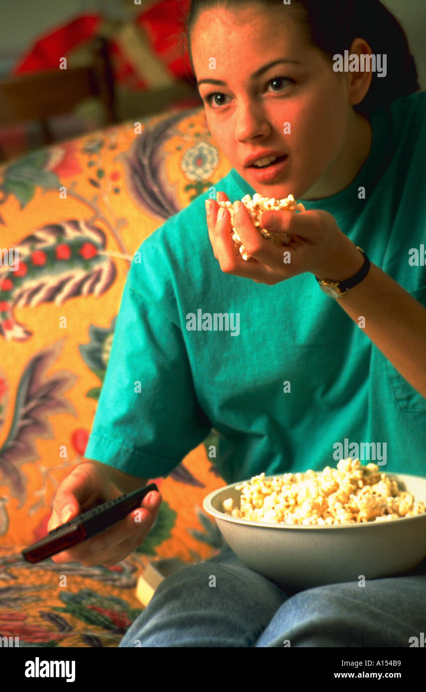 Une adolescente à regarder la télévision sur le canapé eating popcorn avec une main tout en tenant la télécommande de télévision dans l'autre Banque D'Images
