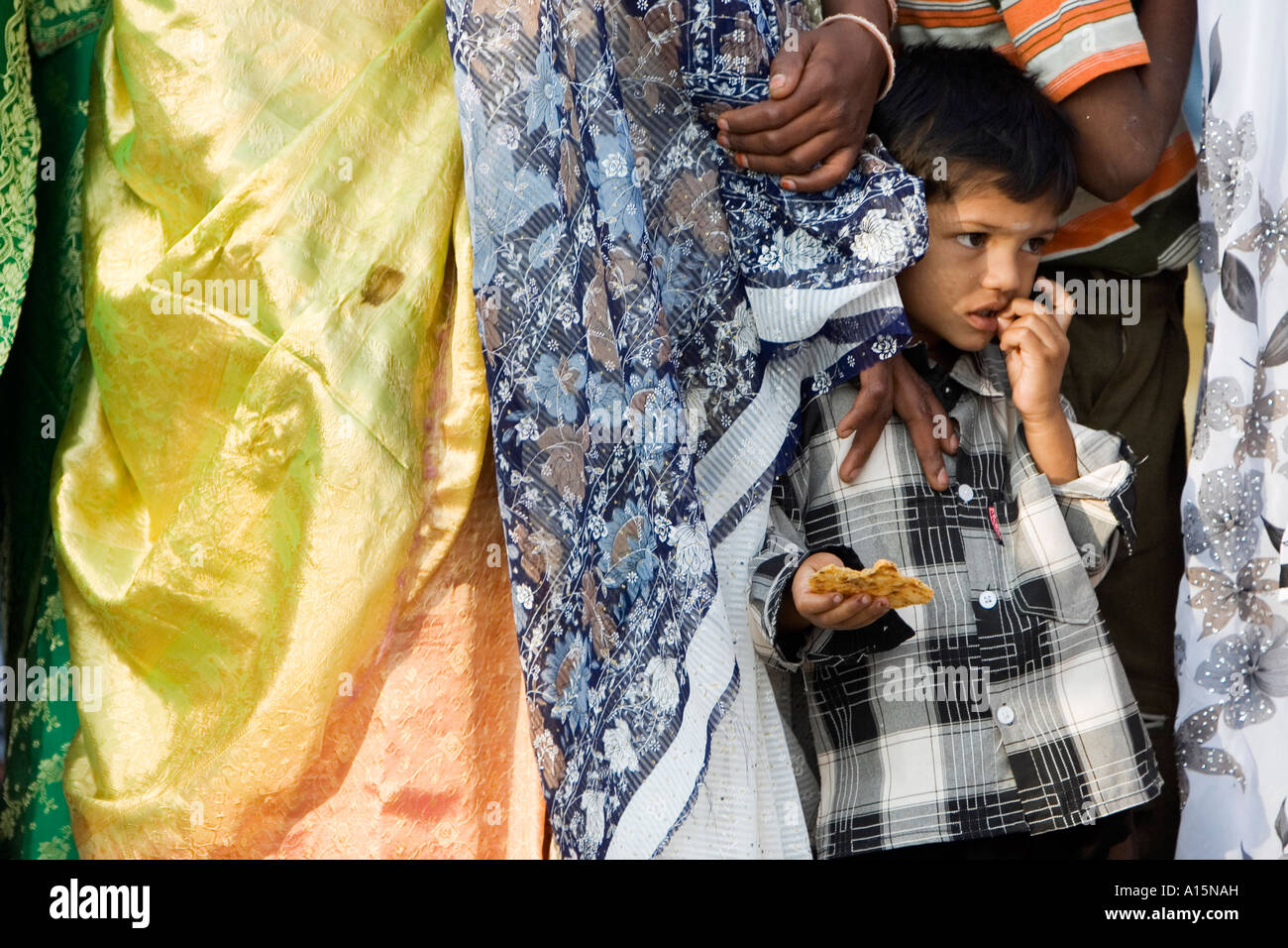 Jeune Indien garçon debout dans une file d'attente entre les femmes portant des saris de manger un biscuit. L'Andhra Pradesh, Inde Banque D'Images