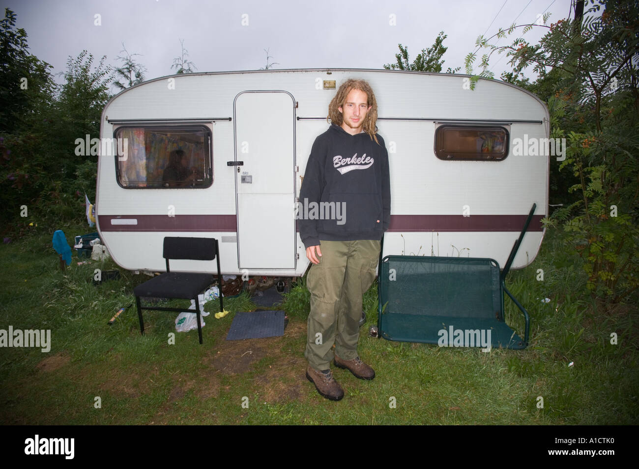 Jeune homme avec des dreadlocks en tenue décontractée, debout devant une caravane dans une plante buissonnante entourant Banque D'Images