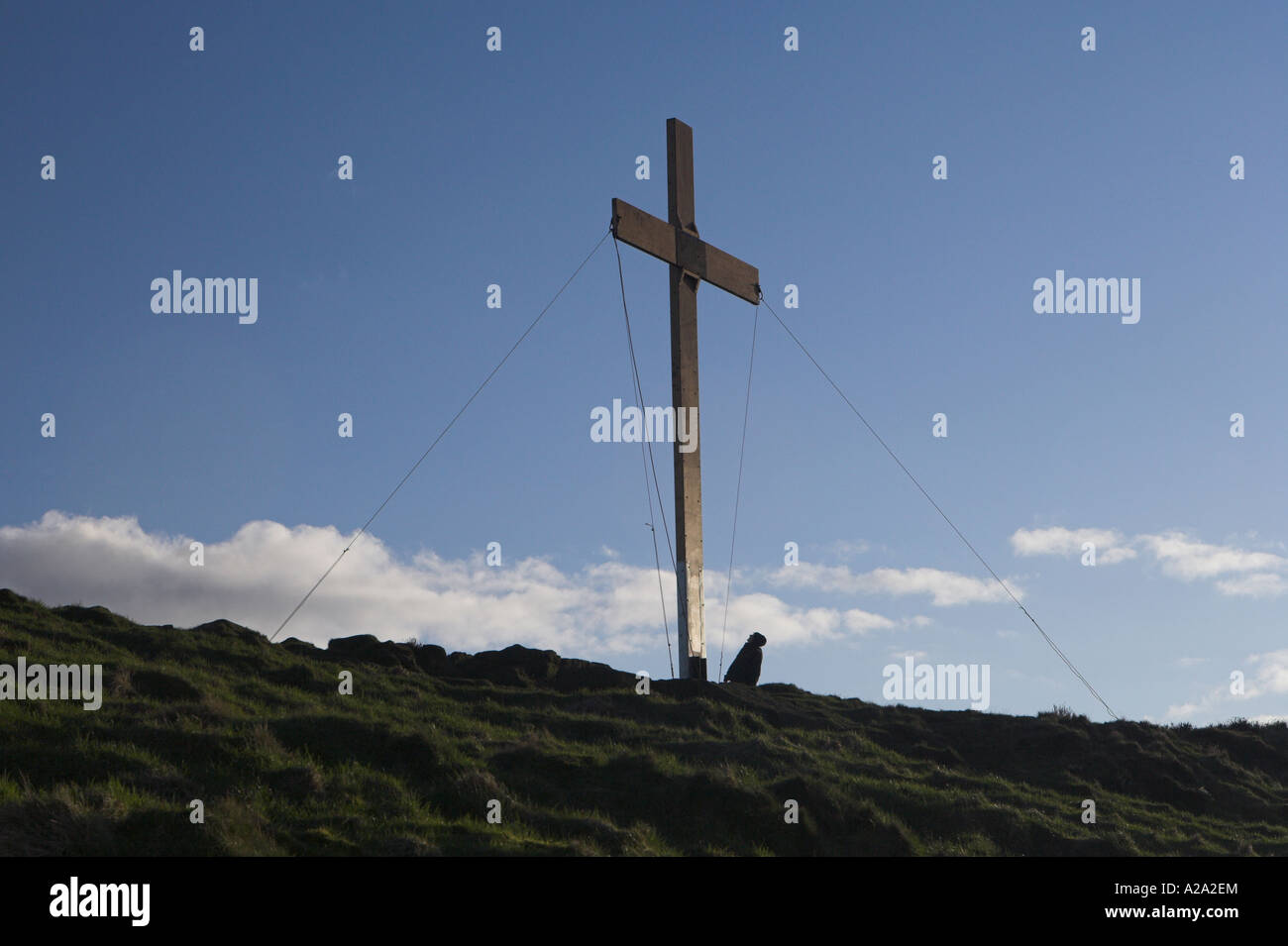 Homme debout au pied d'une grande croix en bois érigée en hauteur sur une colline rurale à Eastertime et dans un ciel bleu profond - Otley Chevin, Yorkshire, Angleterre, Royaume-Uni. Banque D'Images
