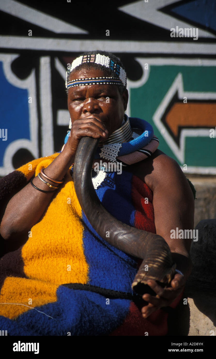 Ndebele woman blowing une corne de kudu en face de doigt painted house, Botshabelo village Ndebele, Afrique du Sud Banque D'Images
