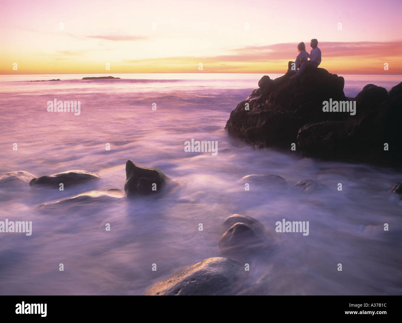 Couple assis sur des roches au coucher du soleil sur la rive peint vague à Laguna Beach, Californie Banque D'Images